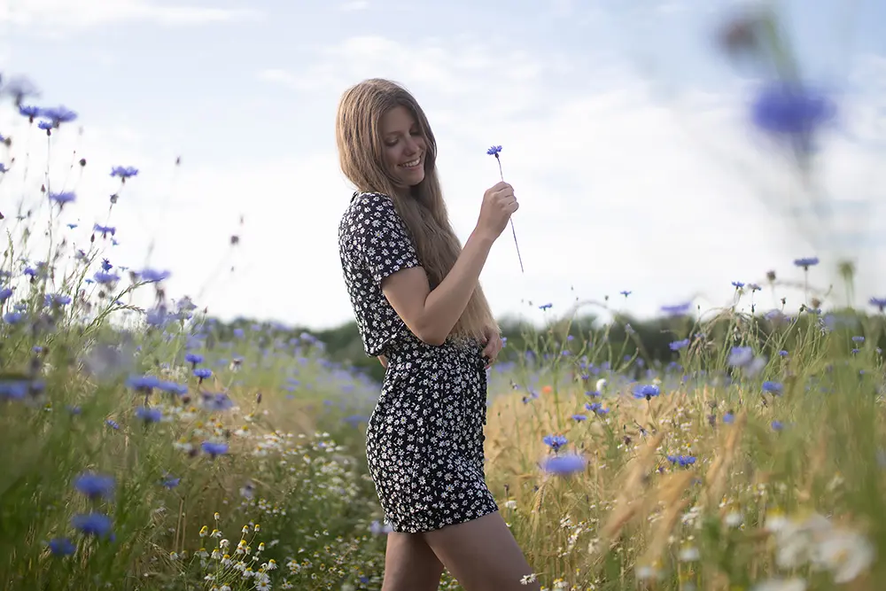 Vanessa steht in einem Feld mit blauen Blüten in einem schwarz-weißen Kleid und hat eine einzelne Blume in der Hand. Das Bild ist unbearbeitet.