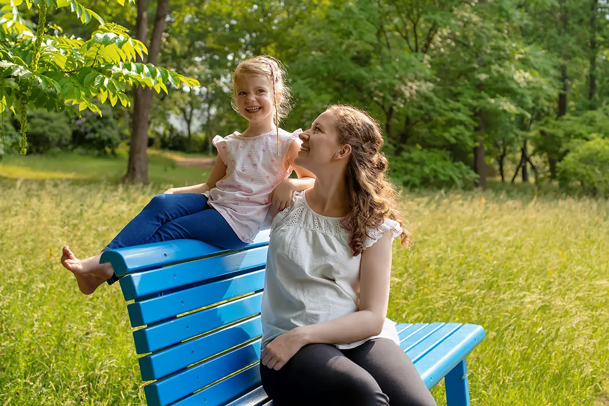 Eine Mutter hält ihre Tochter mit einem Arm fest, wärend die kleine auf der Lehne der blauen Bank sitzt.