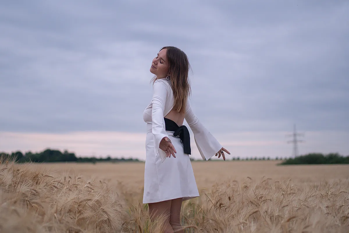 Cora steht im weißen Kleid im Weizenfeld und lächelt mit geschlossenen Augen.